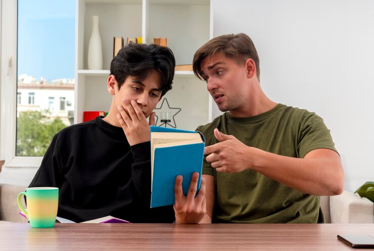 Confident Young Blonde Handsome Man Holding Pointing Book Sitting Table Looking Young Brunette Handsome Guy Putting Hand Mouth Looking Book Inside Living Room 141793 70115